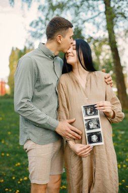 Çiftler çimlerin üzerinde ilk bebeklerini bekleyip kucaklaşırlar. Hamile kadın ve kocası parkta duruyor ve ellerinde bebeğin ultrason fotoğrafı var. Esmer kadın bej elbise giyiyor..