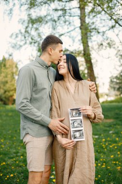 Çiftler çimlerin üzerinde ilk bebeklerini bekleyip kucaklaşırlar. Hamile kadın ve kocası parkta duruyor ve ellerinde bebeğin ultrason fotoğrafı var. Esmer kadın bej elbise giyiyor..