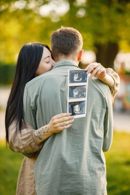 Çiftler çimlerin üzerinde ilk bebeklerini bekleyip kucaklaşırlar. Hamile kadın ve kocası parkta duruyor ve ellerinde bebeğin ultrason fotoğrafı var. Esmer bir kadına odaklan..