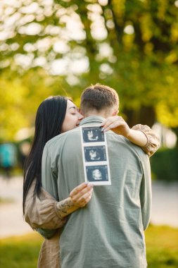 Çiftler çimlerin üzerinde ilk bebeklerini bekleyip kucaklaşırlar. Hamile kadın ve kocası parkta duruyor ve ellerinde bebeğin ultrason fotoğrafı var. Esmer bir kadına odaklan..