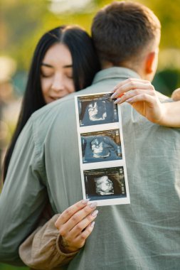 Çiftler çimlerin üzerinde ilk bebeklerini bekleyip kucaklaşırlar. Hamile kadın ve kocası parkta duruyor ve ellerinde bebeğin ultrason fotoğrafı var. Esmer bir kadına odaklan..