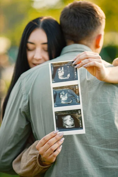 Çiftler çimlerin üzerinde ilk bebeklerini bekleyip kucaklaşırlar. Hamile kadın ve kocası parkta duruyor ve ellerinde bebeğin ultrason fotoğrafı var. Esmer bir kadına odaklan..