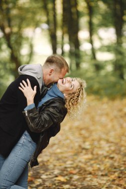 Blonde curly woman and man standing in autumn forest and hugging. Couple wearing leather jackets and blue sweaters. Photo of romantic couple in a forest.