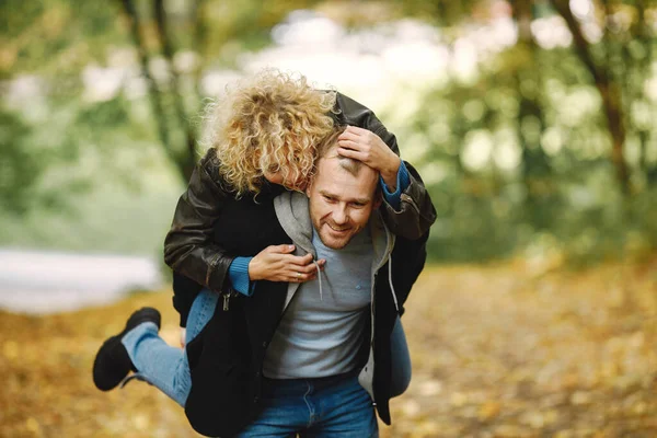Man carrying his woman piggyback in autumn forest. Couple wearing leather jackets and blue sweaters. Photo of romantic couple in a forest.