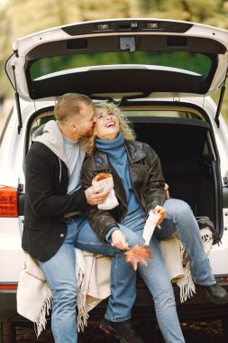 Blonde curly woman and man sitting in a trunk in car in autumn forest and hugging. Couple wearing leather jackets and blue sweaters. Photo of romantic couple in a forest eating a croissants.