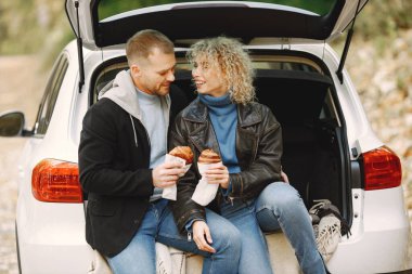 Blonde curly woman and man sitting in a trunk in car in autumn forest and hugging. Couple wearing leather jackets and blue sweaters. Photo of romantic couple in a forest eating a croissants.