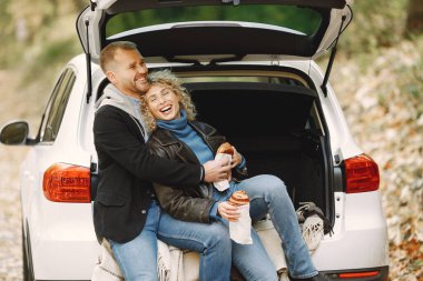 Blonde curly woman and man sitting in a trunk in car in autumn forest and hugging. Couple wearing leather jackets and blue sweaters. Photo of romantic couple in a forest eating a croissants.