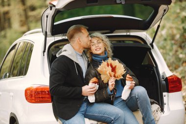 Blonde curly woman and man sitting in a trunk in car in autumn forest and hugging. Couple wearing leather jackets and blue sweaters. Photo of romantic couple in a forest eating a croissants.