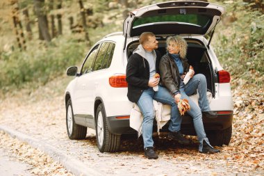 Blonde curly woman and man sitting in a trunk in car in autumn forest and hugging. Couple wearing leather jackets and blue sweaters. Photo of romantic couple in a forest eating a croissants.