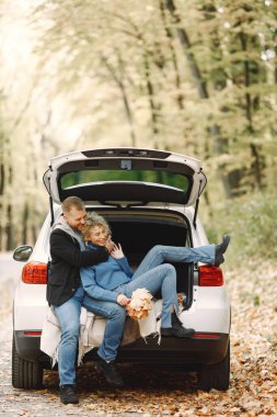 Blonde curly woman and man sitting in a trunk in car in autumn forest and hugging. Man wearing black leather jacket and woman blue sweater. Photo of romantic couple in a forest.