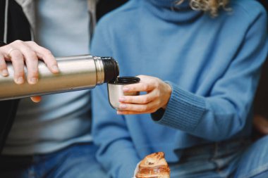 Elinde çay olan termo bardağa odaklan. Kadın ve erkek bagajda oturuyor ve sarılıyorlar. Siyah deri ceket giyen bir adam ve mavi kazak giyen bir kadın..