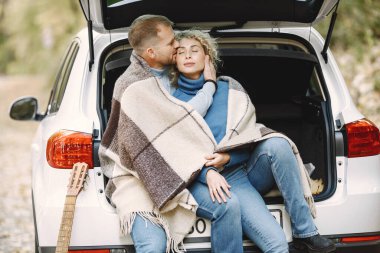 Blonde curly woman and man sitting in a trunk in car in autumn forest with a guitar. Couple wearing leather jackets and blue sweaters. Photo of romantic couple in a forest.