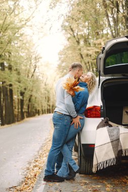 Blonde curly woman and man standing in autumn forest and hugging. Couple wearing blue sweaters. Photo of romantic couple in a forest near an open cars trunk.
