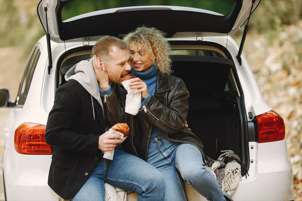 Blonde curly woman and man sitting in a trunk in car in autumn forest and hugging. Couple wearing leather jackets and blue sweaters. Photo of romantic couple in a forest eating a croissants.