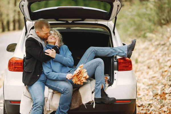 Blonde curly woman and man sitting in a trunk in car in autumn forest and hugging. Man wearing black leather jacket and woman blue sweater. Photo of romantic couple in a forest.