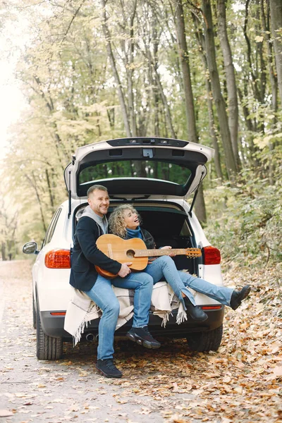 Blonde curly woman and man sitting in a trunk in car in autumn forest with a guitar. Couple wearing leather jackets and blue sweaters. Photo of romantic couple in a forest.