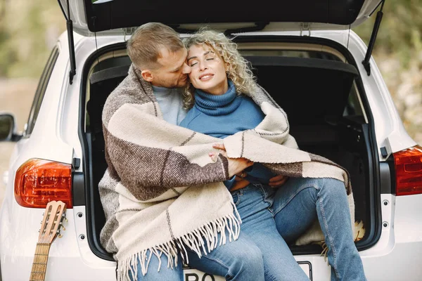 Blonde curly woman and man sitting in a trunk in car in autumn forest with a guitar. Couple wearing leather jackets and blue sweaters. Photo of romantic couple in a forest hugging.
