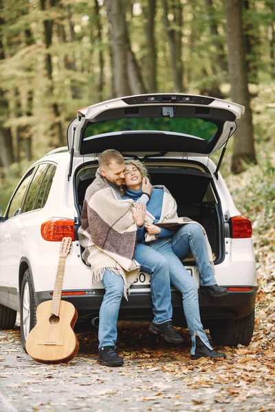 Blonde curly woman and man sitting in a trunk in car in autumn forest with a guitar. Couple wearing leather jackets and blue sweaters. Photo of romantic couple in a forest hugging.