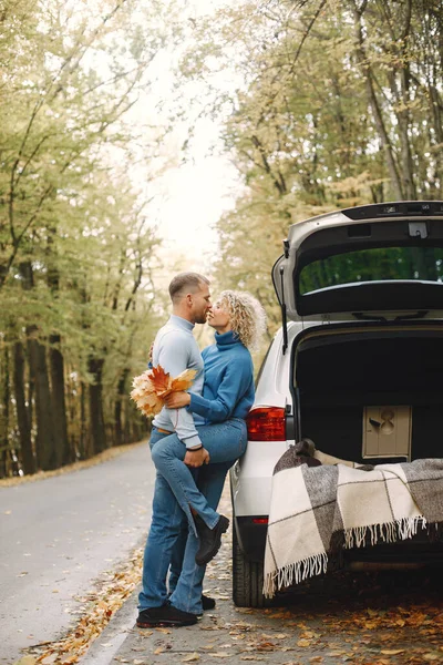 Blonde curly woman and man standing in autumn forest and hugging. Couple wearing blue sweaters. Photo of romantic couple in a forest near an open cars trunk.