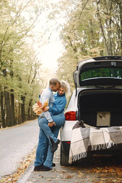 Blonde curly woman and man standing in autumn forest and hugging. Couple wearing blue sweaters. Photo of romantic couple in a forest near an open cars trunk.