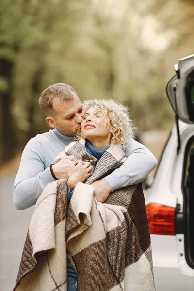 Blonde curly woman and man standing in autumn forest and hugging. Couple wearing blue sweaters. Photo of romantic couple in a forest near an open cars trunk.