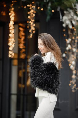 Woman in winter clothes smiles on the background of a cafe with glass doors and Christmas decorations. Brunette woman posing for a photo . Woman wearing black fur coat.