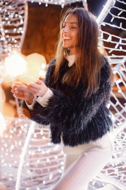 Woman in winter clothes standing near white Christmas lighting in town. Brunette woman holding a sparkler. Woman wearing black fur coat.