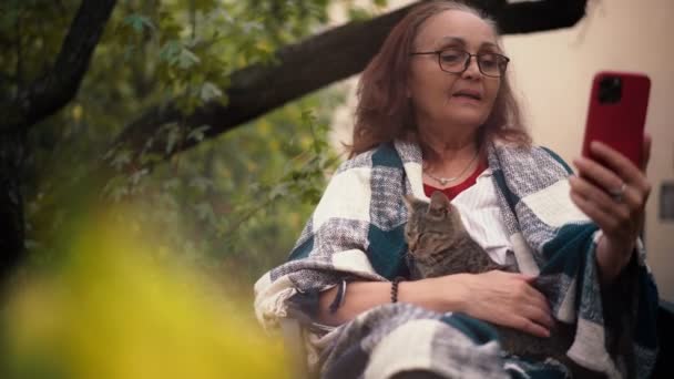 En mogen vuxen kvinna som tar ett videosamtal medan hon sitter i trädgården — Stockvideo
