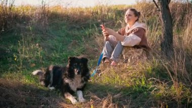 Genç yetişkin bir kadın köpeğiyle yürürken telefonunu kullanıyor.