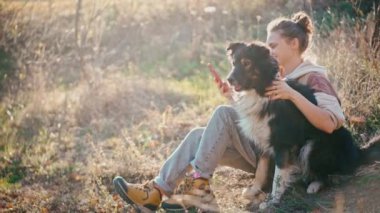 Genç yetişkin bir kadın, bir tepede otururken köpeğiyle selfie çekiyor.