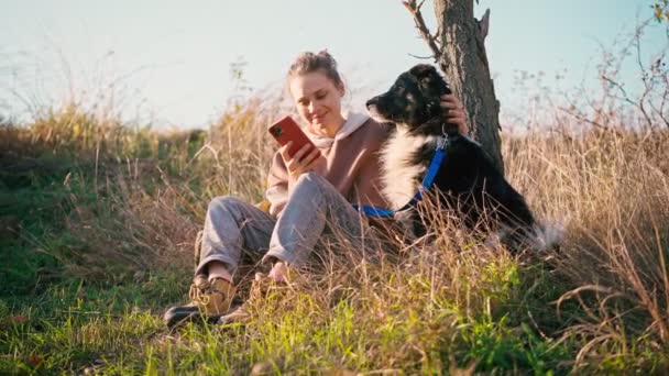 Una mujer joven adulta tomando una selfie con su perro mientras está sentada en una colina — Vídeos de Stock