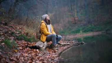 Genç bir kadın bir orman gölünün kıyısında oturuyor ve temiz havanın tadını çıkarıyor..