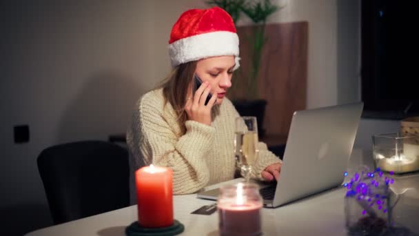 Jonge vrouw met een kerstmuts zit voor een laptop en spreekt aan de telefoon — Stockvideo