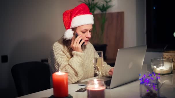 Ung kvinna i en julhatt sitter framför en bärbar dator och talar i telefon — Stockvideo