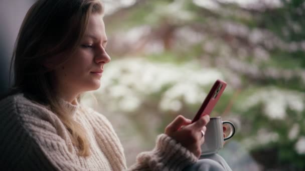 Una joven se sienta en el alféizar de la ventana y bebe café mientras usa su teléfono inteligente — Vídeos de Stock