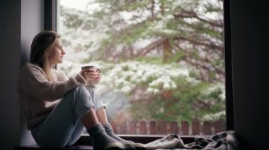 Güzel genç bir kız pencere eşiğinde oturur ve fincandan kahve içer.