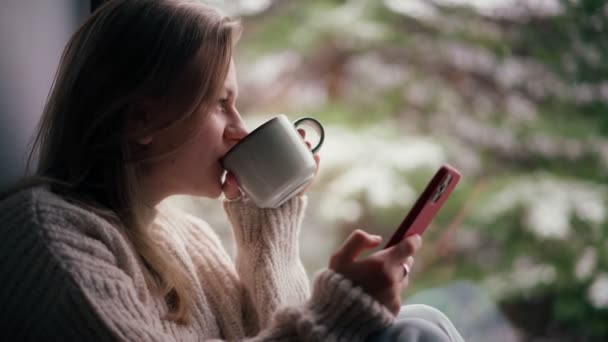Młoda dziewczyna siedzi na parapecie i pije kawę podczas korzystania ze smartfona — Wideo stockowe