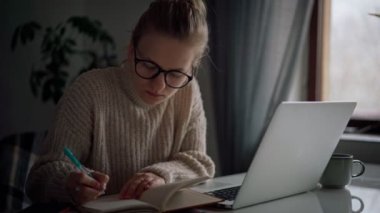 Dizüstü bilgisayarda yazan ve not defterine not alan gözlüklü genç bir kız.