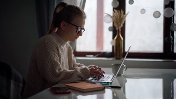 Una joven con gafas escribiendo en un portátil y tomando notas en un cuaderno — Vídeos de Stock
