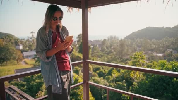 A young adult woman using her smartphone while standing on a terrace with a view — Stock Video