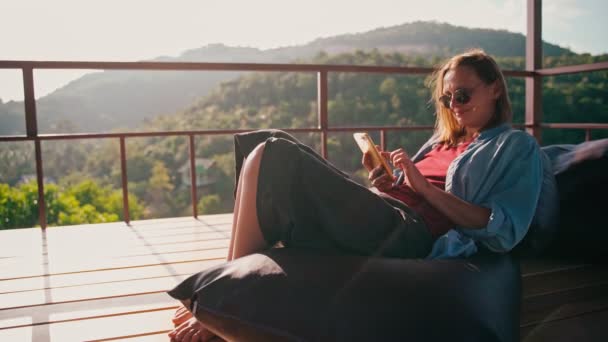 Una joven mujer alegre adulta usando su teléfono inteligente mientras está sentada en una silla de bolsa — Vídeo de stock