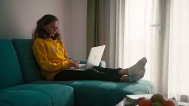 A young adult woman coughs while sitting on the couch and working on a laptop