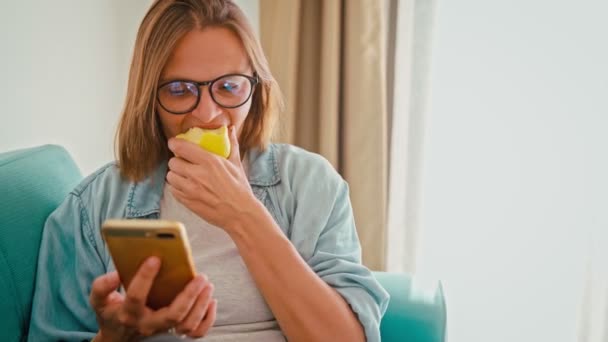 Young adult woman wearing glasses eating an apple while holding a smartphone — Stockvideo
