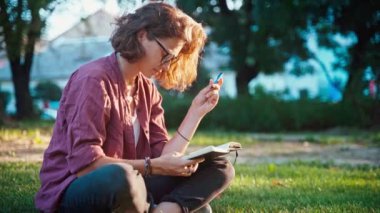 A young woman sitting on the lawn in the park and making notes in her notebook.