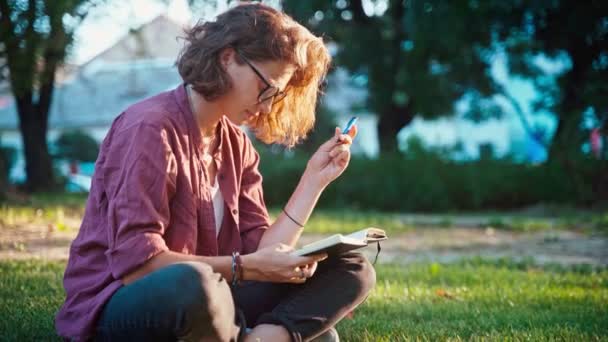 Una joven sentada en el césped del parque y anotando en su cuaderno. — Vídeos de Stock