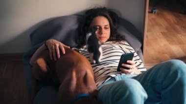 Genç bir kadın, büyük bir yastıkta uzanmış köpeğine ve küçük bir kediye sarılırken elinde telefonla rahatlar.