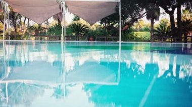 El yapımı sinemasal bir çekim. Güneş yatakları ve palmiye ağaçlarının arka planında su yüzeyinde küçük dalgalanmalar var..