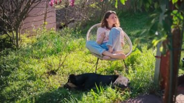 Manolya ağacının altındaki bahçede bir sandalyede oturmuş sabah kahvesinin tadını çıkaran güzel bir kadın ve köpeği etrafta geziniyor..