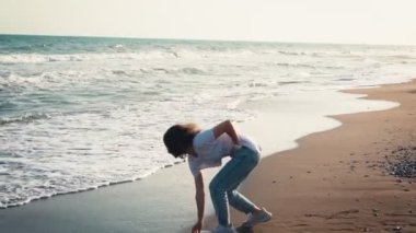 Gün batımında kıyıda yürürken deniz dalgalarına taş atan genç bir kadın.