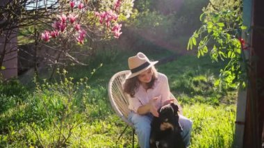 Genç ve güzel bir kadın bir kır evinin arka bahçesinde sandalyede otururken sevimli köpeğiyle oynuyor..